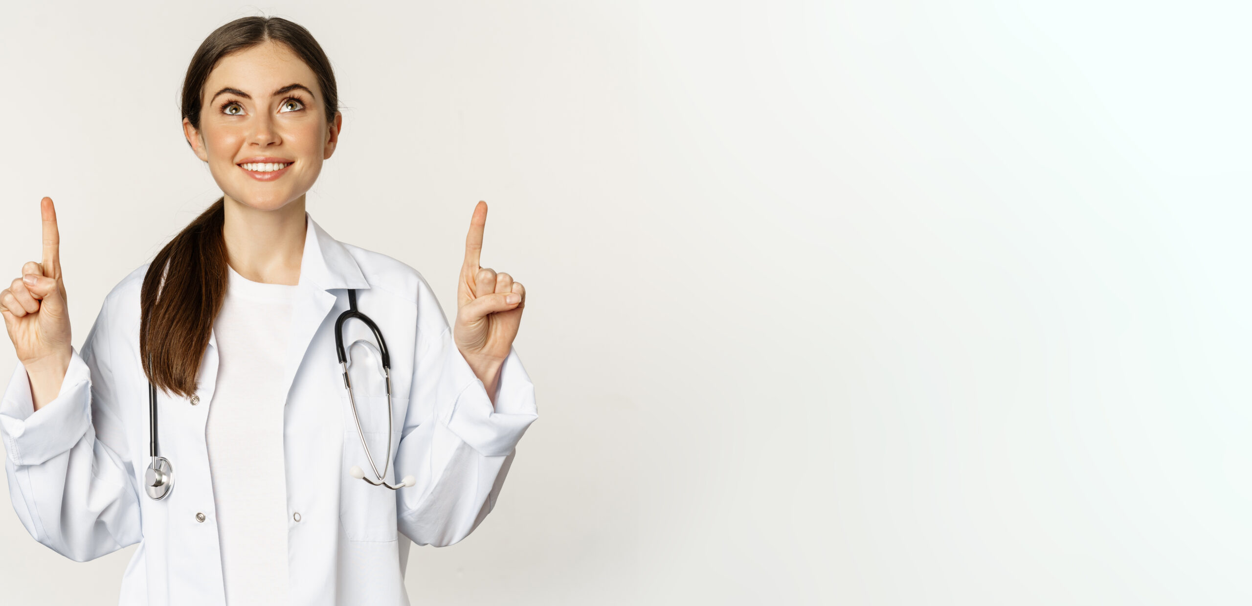 Happy beautiful doctor, female physician pointing fingers up, showing advertisement, standing over white background. Copy space